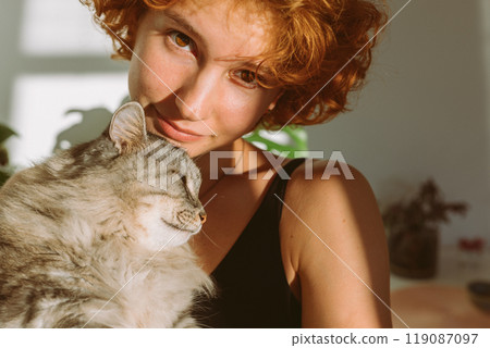 portrait red-haired curly young woman with pet gray fluffy cat in arms, lit by bright sun 119087097