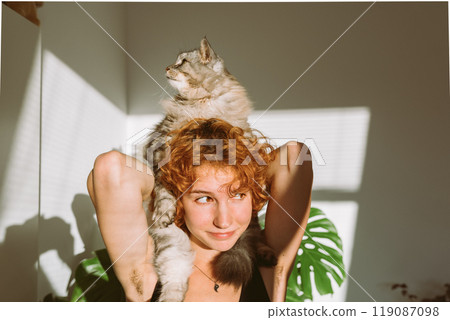 portrait red-haired curly young woman with pet gray fluffy cat in arms, lit by bright sun 119087098