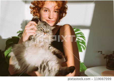 portrait red-haired curly young woman with pet gray fluffy cat in arms, lit by bright sun portrait red-haired curly young woman with pet gray fluffy cat in arms, lit by bright sun 119087099