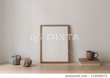Autumn interior still life. Blank wooden photo frame mockup on table with pumpkins. Thanksgiving, Halloween home decor. Scandi interior. Cup of tea, coffee on books. White wall background 119090678