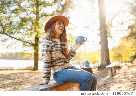 Woman holds thermos and drinks tea in sunny autumn forest. Vacation, travel, lifestyle concept. 119090707