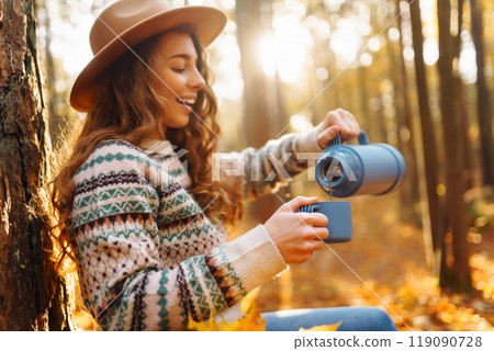 Cute young woman drinks hot drink from thermos and enjoys nature. Autumn landscape. relax concept. 119090728