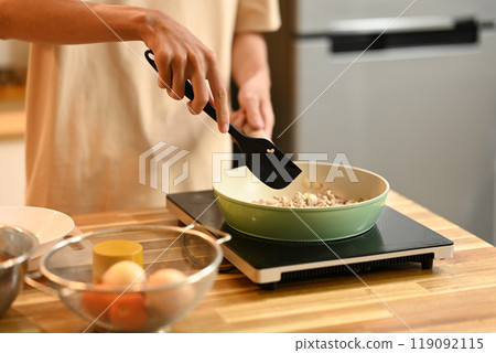 Close up shot of young man cooking, stirring ingredients in frying pan with a spatula 119092115