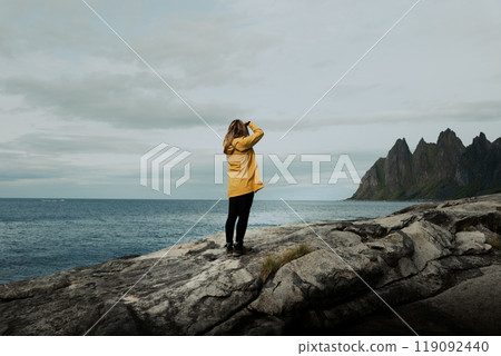 Female in a Yellow Jacket shoots seascape with a camera with mountains in background Female in a Yellow Jacket shoots seascape with a camera with mountains in background 119092440