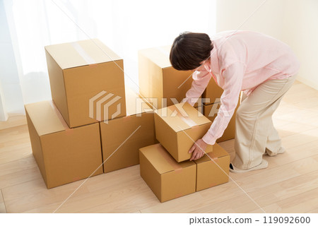 A middle-aged woman carrying many cardboard boxes containing moving luggage into a room 119092600