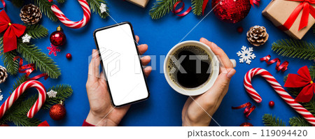 Above view of female hand holding smart phone with hot cup of coffee. Christmas decoration on colored background Above view of female hand holding smart phone with hot cup of coffee. Christmas decoration on colored background 119094420