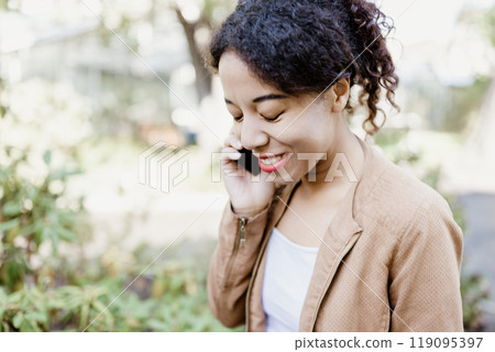 Young Multiracial Woman Talking on Smartphone Outdoors in Relaxed Setting, smiling 119095397
