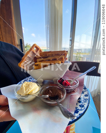 Close-up male holding a plate of waffles with sides of ice cream, berries, and chocolate at a seaside restaurant. Bright and sunny setting, with palm trees visible outside, creates a relaxed vacation Close-up male holding a plate of waffles with sides of ice cream, berries, and chocolate at a seaside restaurant. Bright and sunny setting, with palm trees visible outside, creates a relaxed vacation 119096648