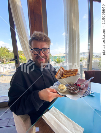 Man smiles while holding a plate of waffles with sides of ice cream, berries, and chocolate at a seaside restaurant. Bright and sunny setting, with palm trees visible outside, creates a relaxed Man smiles while holding a plate of waffles with sides of ice cream, berries, and chocolate at a seaside restaurant. Bright and sunny setting, with palm trees visible outside, creates a relaxed 119096649