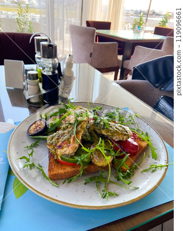 Close-up plate of grilled vegetables and chicken on toast is served in a stylish, bright seaside restaurant. Dish is garnished with fresh arugula and accompanied by a variety of seasonings and 119096661