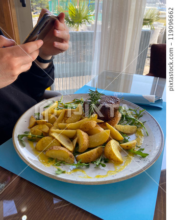 Close-up man takes a photo of a dish consisting of grilled meat and potatoes. Social networks, blog and food blogger concept 119096662