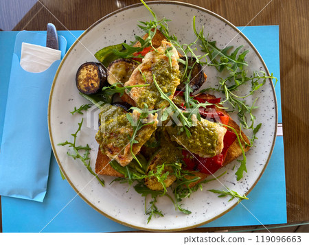 Close-up plate of grilled vegetables and chicken on toast is served in a stylish, bright seaside restaurant. Dish is garnished with fresh arugula and accompanied by a variety of seasonings and Close-up plate of grilled vegetables and chicken on toast is served in a stylish, bright seaside restaurant. Dish is garnished with fresh arugula and accompanied by a variety of seasonings and 119096663