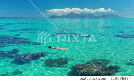 Woman is snorkeling in a shallow lagoon with turquoise water and a mountainous island in the background. The water is so clear that the coral reef is visible 119096884