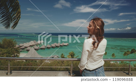 Young woman is taking in the stunning panoramic view of turquoise waters, luxurious overwater bungalows, and lush greenery 119096888