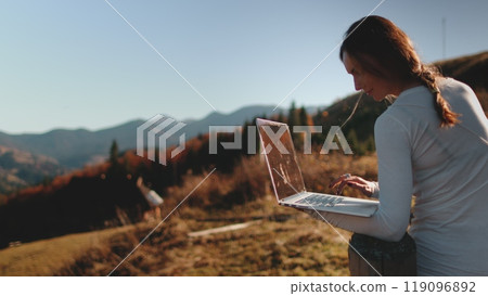 A confident woman stands atop a hill, her eyes fixed on the laptop screen in her hands. She seems both connected to the digital world and in tune with the natural beauty surrounding her. 119096892