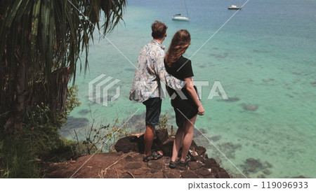 Young couple is embracing on a cliff edge while looking out over a beautiful tropical ocean with sailboats in the distance 119096933
