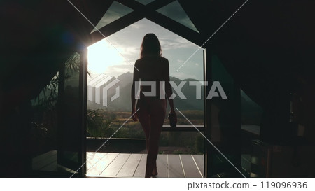 Woman is exiting a luxury bungalow and walking towards a breathtaking mountain view at sunrise, starting her day with peace and tranquility 119096936