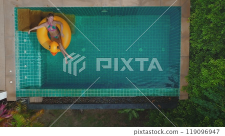 Aerial view woman relaxing on yellow duck float in a swimming pool. The pool is surrounded by green foliage and the water is clear and blue. The woman in bikini and is enjoying the sunshine. Aerial view woman relaxing on yellow duck float in a swimming pool. The pool is surrounded by green foliage and the water is clear and blue. The woman in bikini and is enjoying the sunshine. 119096947