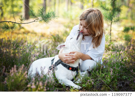 A girl shares a loving kiss with her white dog in a sunlit forest during a peaceful afternoon A girl shares a loving kiss with her white dog in a sunlit forest during a peaceful afternoon 119097377