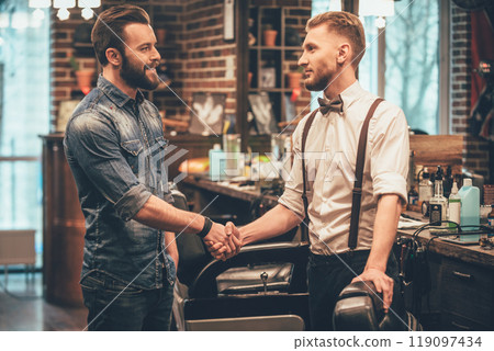 Thank you for quality haircut! Barber and his client shaking hands with smile while standing at barbershop 119097434