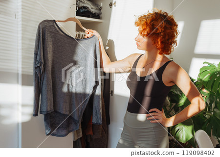 portrait of young red-haired woman choosing clothes in closet at home 119098402