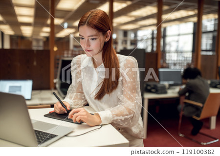 Long-haired pretty ginger girl working on a laptop in the library 119100382