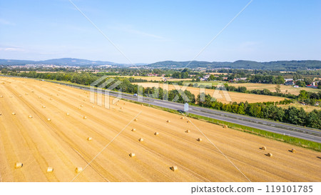 Aerial perspective showing a field of hay bales beside a highway, surrounded by green trees and distant hills under a clear blue sky. 119101785