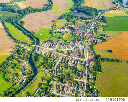 The vibrant rural landscape of Trebonsko showcases a picturesque layout of fields and houses, interspersed with winding roads and a serene river. Green and golden hues paint the idyllic scene. 119101790