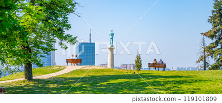 Visitors relax on benches near the Victor Monument, overlooking the expansive views from Kalemegdan Fortress, a historic site in Belgrade, Serbia, under a clear blue sky. 119101809