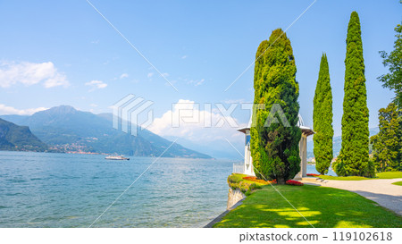 A tranquil lakeside scene in Bellagio, Italy, showcases lush greenery and tall trees along the shore of Como Lake, with distant mountains and a boat sailing in the serene waters. 119102618