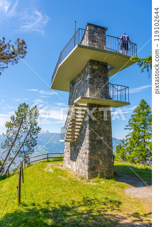 A visitor explores the Gaflei lookout tower in Liechtenstein, enjoying panoramic views of the surrounding mountains and landscape under a clear blue sky. A visitor explores the Gaflei lookout tower in Liechtenstein, enjoying panoramic views of the surrounding mountains and landscape under a clear blue sky. 119102644