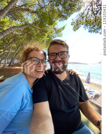 Couple man and woman at the beach under a tree in a sunny day taking selfie for social networks and travel blog. Blogger and journey concept Couple man and woman at the beach under a tree in a sunny day taking selfie for social networks and travel blog. Blogger and journey concept 119103431