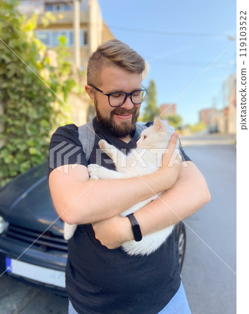 Bearded man gently embraces a white cat, conveying a moment of warmth and connection with pet. Outdoor setting adds an element of everyday life and companionship Bearded man gently embraces a white cat, conveying a moment of warmth and connection with pet. Outdoor setting adds an element of everyday life and companionship 119103522