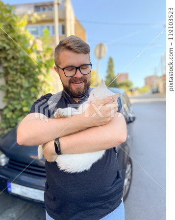 Bearded man gently embraces a white cat, conveying a moment of warmth and connection with pet. Outdoor setting adds an element of everyday life and companionship 119103523