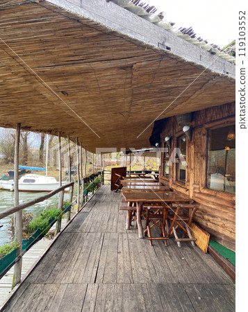 Rustic waterfront deck with wooden tables and a thatched roof overlooks serene river. Outdoor seating area offers a tranquil place to relax and enjoy the natural surroundings 119103552