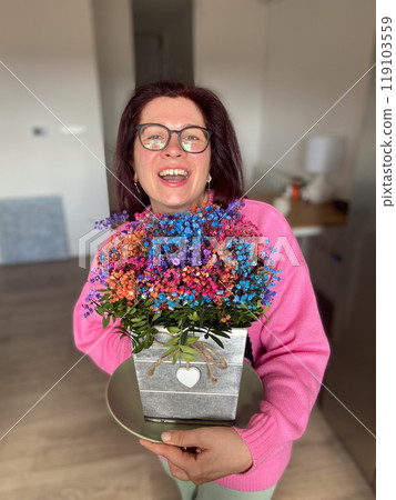 Cheerful millennial woman with glasses is joyfully holding a colorful bouquet of flowers in a decorative pot. Pink sweater and radiant smile add warmth and vibrancy to the scene 119103559