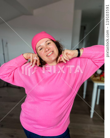 Millennial woman joyfully poses in a bright pink sweater and matching hat, exuding happiness and confidence in a casual home setting. Cheerful demeanor and vibrant outfit create an uplifting and 119103591