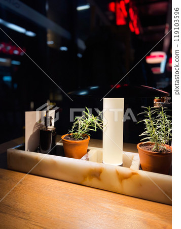 Cozy restaurant table setting with olive oil and balsamic vinegar bottles, flanked by small potted rosemary plants under warm lighting. Blurred background evokes a bustling yet intimate atmosphere 119103596