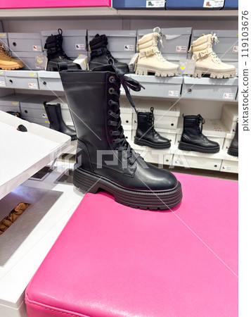 Black leather combat boot displayed on a bright pink bench in a shoe store. Various other boots in different colors and styles are neatly stacked in the background Black leather combat boot displayed on a bright pink bench in a shoe store. Various other boots in different colors and styles are neatly stacked in the background 119103676