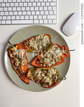 Delicious stuffed peppers with melted cheese and herbs on a plate, placed near a computer keyboard and mouse on a white desk. Perfect blend of work and healthy eating 119103680
