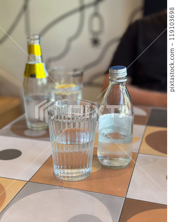 Glass of water alongside a bottle on a patterned tile table. Depicting everyday hydration and minimalistic lifestyle themes. Glass of water alongside a bottle on a patterned tile table. Depicting everyday hydration and minimalistic lifestyle themes. 119103698