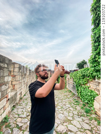 A man takes a photo while standing at a viewpoint in fortress. A tourist with a smartphone camera A man takes a photo while standing at a viewpoint in fortress. A tourist with a smartphone camera 119103722