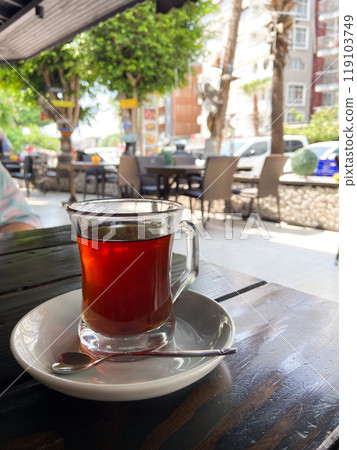 Glass of tea on a white saucer with a spoon, set on an outdoor cafe table with a view of a bustling urban street scene in the background. Essence of a relaxing tea moment amidst city life 119103749