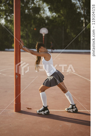A lively and energetic skater is seen stretching at an outdoor basketball court, 119104158