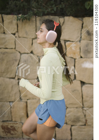 A motivated and active woman is jogging outdoors while wearing her headphones, enjoying her workout 119104300