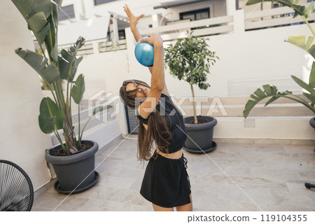 A Joyful Young Woman Stretching with an Exercise Ball in a Beautiful Outdoor Setting 119104355