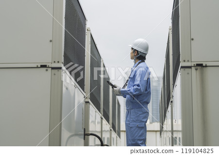 Building maintenance image: A man in work clothes inspecting the outdoor air conditioning unit installed on the roof of a building 119104825