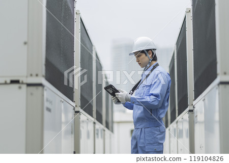Building maintenance image: Profile of a man in work clothes inspecting an air conditioning unit installed on the roof of a building 119104826