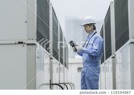 Building maintenance image: A man in work clothes inspecting the outdoor air conditioning unit installed on the roof of a building 119104867