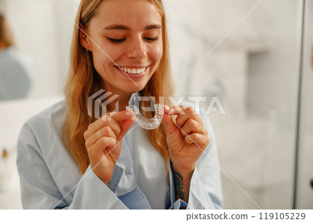 A Smiling Woman Holding a Clear Orthodontic Aligner While Standing in Her Bathroom Setting A Smiling Woman Holding a Clear Orthodontic Aligner While Standing in Her Bathroom Setting 119105229